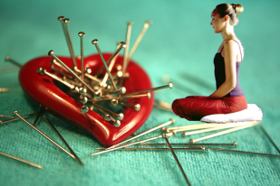 Miniature figue meditating on  a heart shaped pin cushion full of pins 